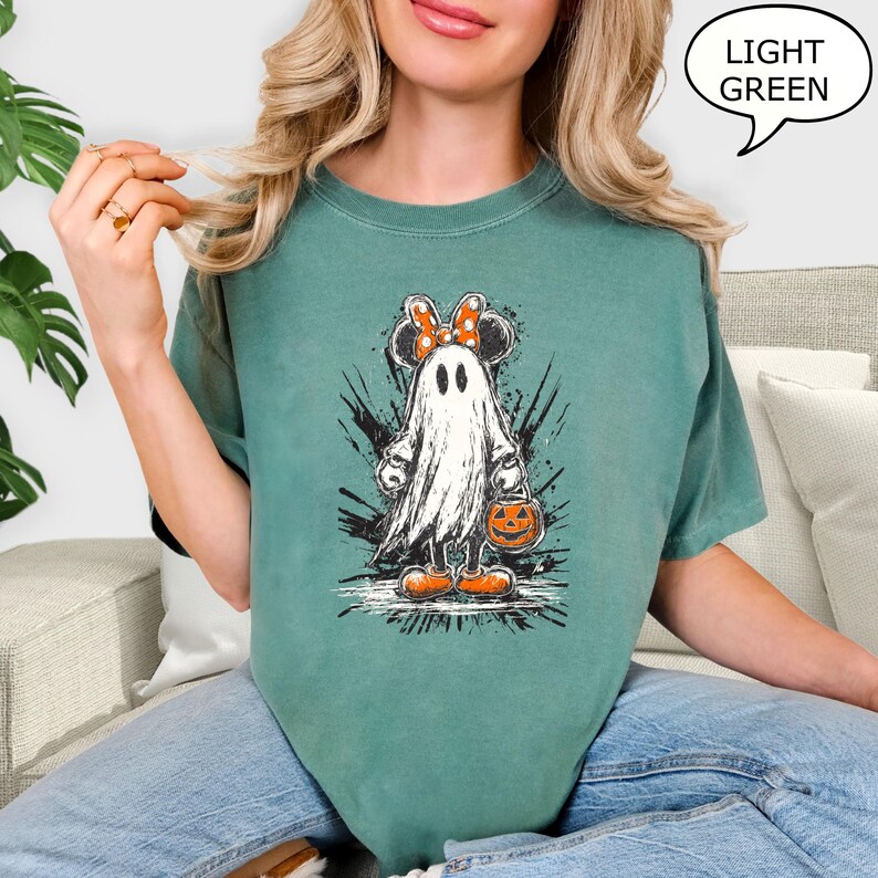 May include: A light green t-shirt featuring a cartoon ghost with Minnie Mouse ears and a bow. The ghost is holding a jack-o'-lantern bucket. The shirt has the text "LIGHT GREEN" in a speech bubble.