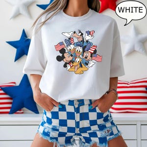 May include: White t-shirt featuring a graphic of Mickey Mouse, Donald Duck, Goofy, and Pluto, each holding American flags or wearing patriotic accessories. The word "WHITE" is in a speech bubble. The shirt is paired with blue and white checkered shorts.