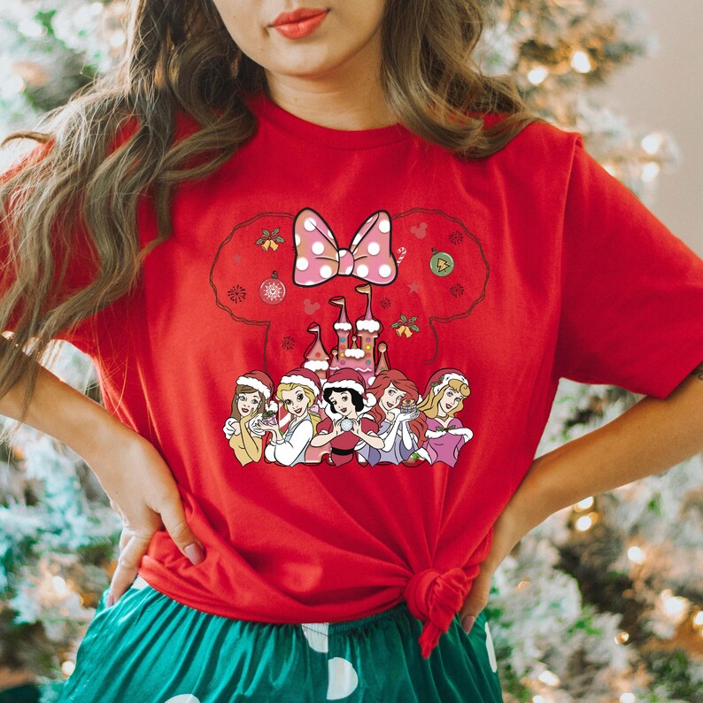 Puede incluir: Camiseta roja con un dise&ntilde;o navide&ntilde;o con una imagen de dibujos animados de un castillo, un lazo y seis princesas Disney con gorros de Pap&aacute; Noel. Las princesas son Blancanieves, Cenicienta, Ariel, Bella, Aurora y Rapunzel.