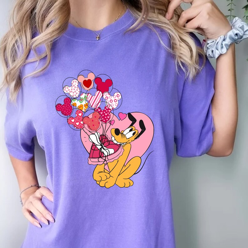 Może przedstawiać: Fioletowy T-shirt z kresk&oacute;wkowym obrazkiem Plutona trzymającego pudełko czekoladek w kształcie serca i otoczonego balonami w kształcie serca.