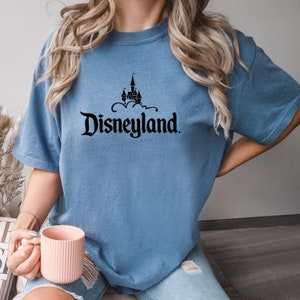 Puede incluir: Camiseta azul claro con el logotipo negro de Disneyland. El logotipo muestra un castillo sobre la palabra "Disneyland". La camiseta tiene mangas cortas y un corte holgado.