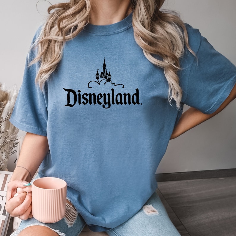 Puede incluir: Camiseta azul claro con el logotipo negro de Disneyland. El logotipo muestra un castillo sobre la palabra "Disneyland". La camiseta tiene mangas cortas y un corte holgado.