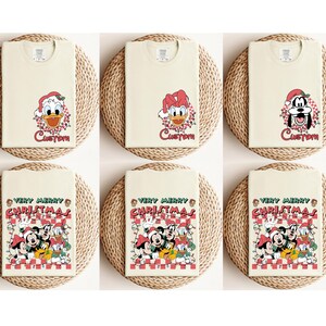 May include: Six cream-colored t-shirts with Christmas-themed graphics. The top row features Donald Duck and Goofy in Santa hats with the word "Custom". The bottom row displays Mickey, Minnie, Donald, and Goofy with the text "Very Merry Christmas".