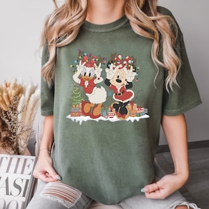Puede incluir: Una camiseta verde con un gr&aacute;fico navide&ntilde;o que presenta a Minnie Mouse y Daisy Duck con gorros de Pap&aacute; Noel y levantando las manos en se&ntilde;al de paz. El gr&aacute;fico tambi&eacute;n incluye un &aacute;rbol de Navidad, regalos y luces.