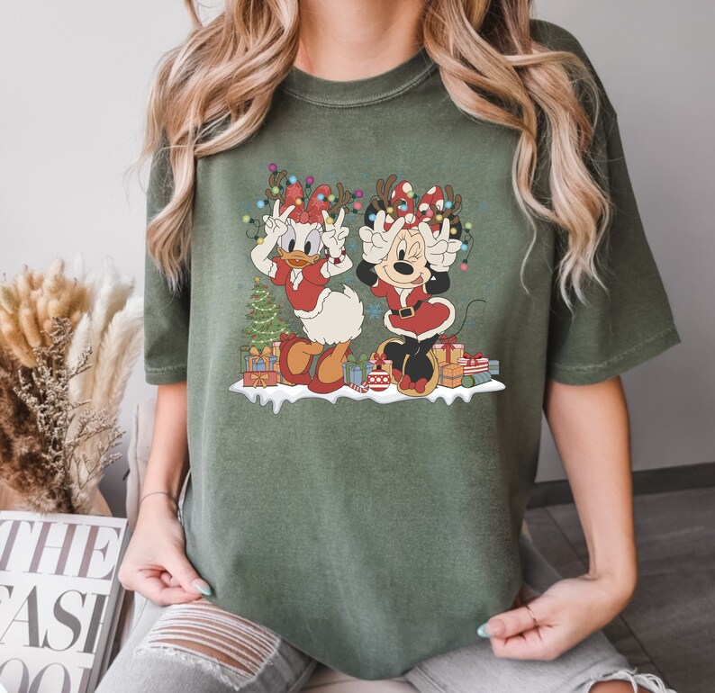 Puede incluir: Una camiseta verde con un gr&aacute;fico navide&ntilde;o que presenta a Minnie Mouse y Daisy Duck con gorros de Pap&aacute; Noel y levantando las manos en se&ntilde;al de paz. El gr&aacute;fico tambi&eacute;n incluye un &aacute;rbol de Navidad, regalos y luces.