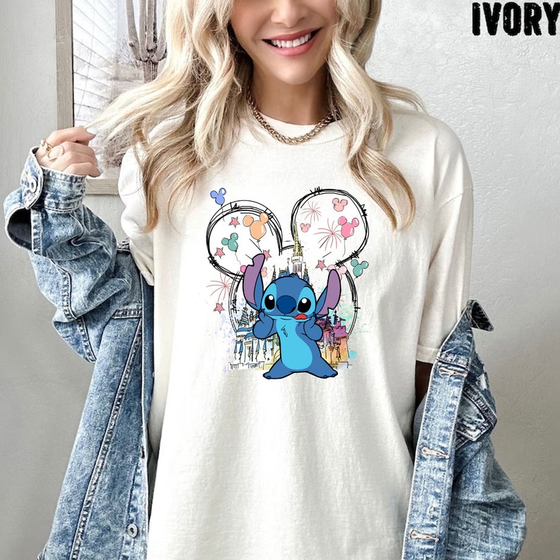 May include: A white t-shirt with a blue Stitch character holding balloons in front of a cartoon drawing of Cinderella's castle. The balloons are pink, blue, yellow, and green.