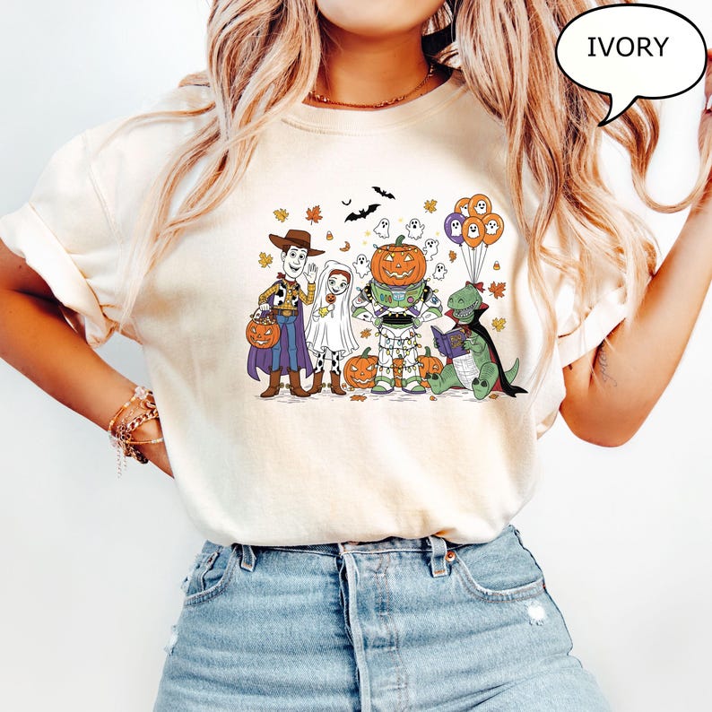 Puede incluir: Camiseta de color marfil con un gr&aacute;fico de Halloween. El dise&ntilde;o presenta personajes de dibujos animados disfrazados, incluyendo a Woody, Buzz Lightyear y un dinosaurio leyendo un libro. La camiseta incluye la palabra "IVORY" en una burbuja.