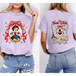 May include: A light purple t-shirt featuring a Snow White graphic on the front and back. The front design includes Snow White holding an apple within a gold frame with a red bow. The back design features the words "Snow White" and other characters.