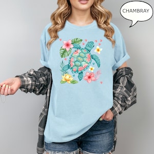 May include: Light blue t-shirt with a watercolor design of a sea turtle, surrounded by tropical flowers and leaves. The turtle has a teal shell with pink floral accents. The word "CHAMBRAY" is in a speech bubble.