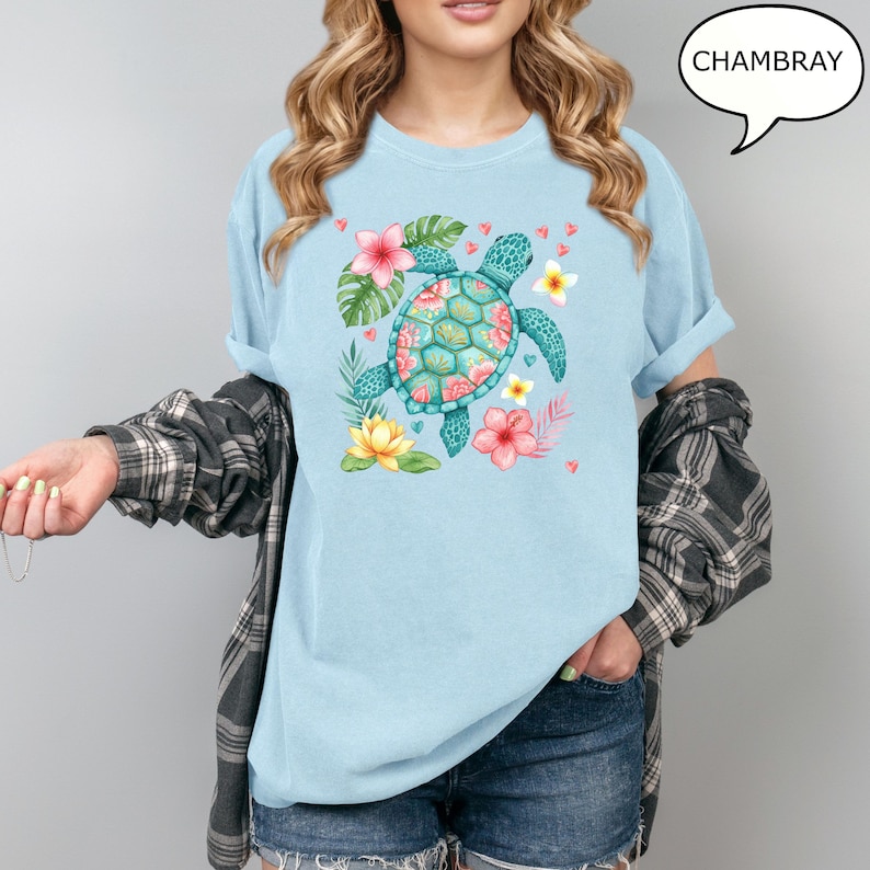 May include: Light blue t-shirt with a watercolor design of a sea turtle, surrounded by tropical flowers and leaves. The turtle has a teal shell with pink floral accents. The word "CHAMBRAY" is in a speech bubble.