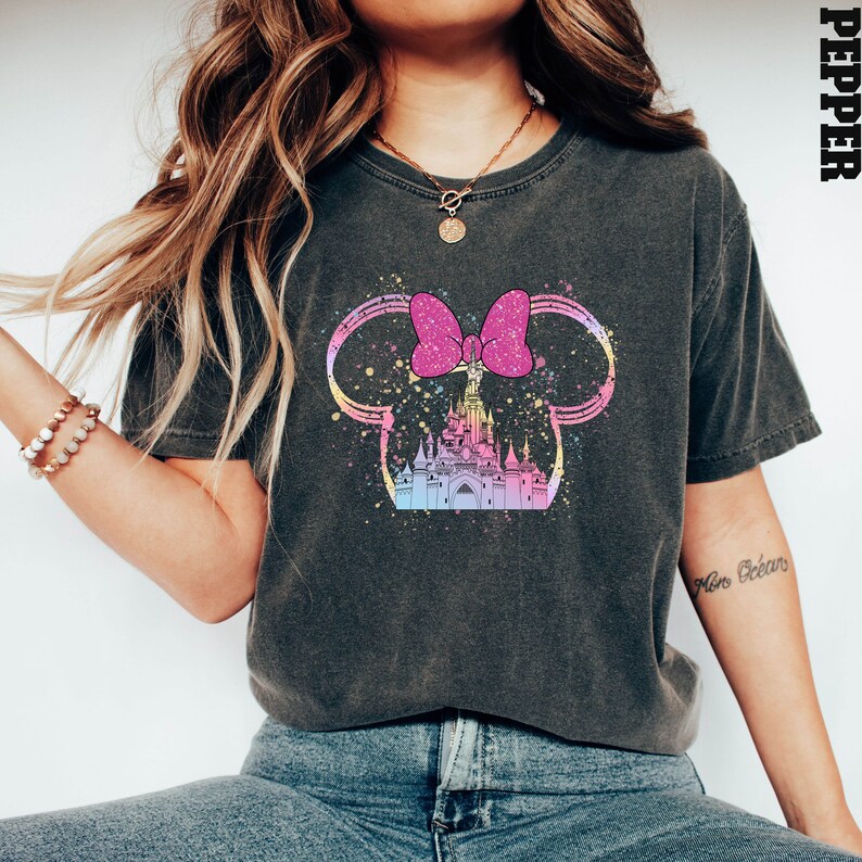 Puede incluir: Una camiseta negra con un gr&aacute;fico de un lazo rosa y un castillo en un esquema de colores arco&iacute;ris. El gr&aacute;fico tiene la forma de las orejas de Mickey Mouse.