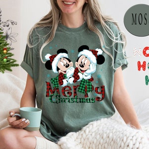 May include: Green t-shirt with a Christmas-themed design featuring Mickey and Minnie Mouse wearing Santa hats and scarves. The text "Merry Christmas" is written in red and black plaid.