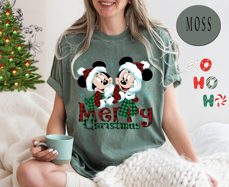 May include: Green t-shirt with a Christmas-themed design featuring Mickey and Minnie Mouse wearing Santa hats and scarves. The text "Merry Christmas" is written in red and black plaid.