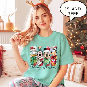 Puede incluir: Una camiseta verde claro con cuatro personajes de dibujos animados con atuendos navide&ntilde;os. Los personajes son el Pato Donald, Minnie Mouse, Mickey Mouse y Goofy. Las palabras "Merry Christmas" est&aacute;n escritas en cursiva debajo. Un bocadillo dice "ISLAND REEF".