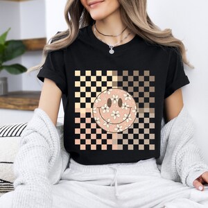 Peut inclure: T-shirt noir avec un motif &agrave; carreaux beige et marron. Un smiley rose avec des fleurs blanches est centr&eacute; sur le motif &agrave; carreaux.