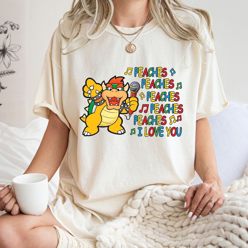 May include: Cream-colored t-shirt featuring a cartoon Bowser character holding a microphone. The text "PEACHES" is repeated multiple times in various colors, along with musical notes and the phrase "I LOVE YOU".