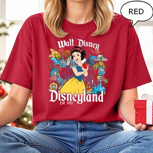 May include: Red t-shirt with a graphic of Snow White holding an apple, and the text "Walt Disney Disneyland Est. 1955". The design includes Disney park icons. The shirt is a solid red colour.