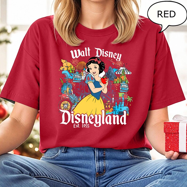 May include: Red t-shirt with a graphic of Snow White holding an apple, and the text "Walt Disney Disneyland Est. 1955". The design includes Disney park icons. The shirt is a solid red colour.