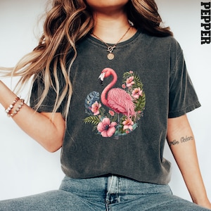 K&ouml;nnte beinhalten: Ein dunkelgraues T-Shirt mit einem pinkfarbenen Flamingo und tropischen Blumen, die auf der Vorderseite aufgedruckt sind. Das Shirt hat eine lockere Passform und einen Rundhalsausschnitt.