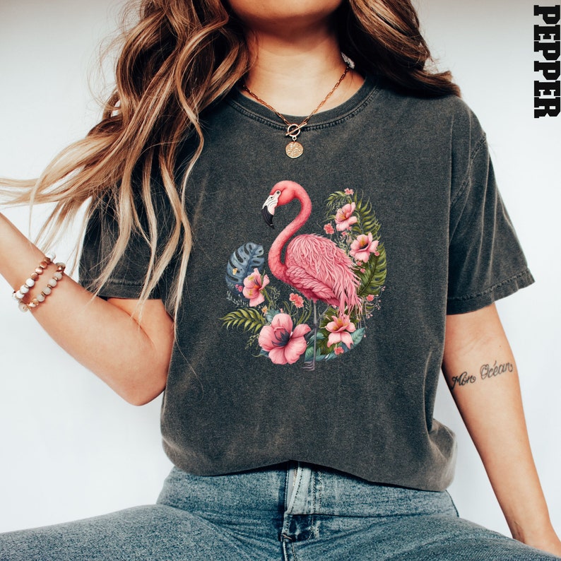K&ouml;nnte beinhalten: Ein dunkelgraues T-Shirt mit einem pinkfarbenen Flamingo und tropischen Blumen, die auf der Vorderseite aufgedruckt sind. Das Shirt hat eine lockere Passform und einen Rundhalsausschnitt.