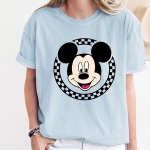 Puede incluir: Camiseta azul claro con un c&iacute;rculo a cuadros blanco y negro con una imagen de dibujos animados de la cara de Mickey Mouse.