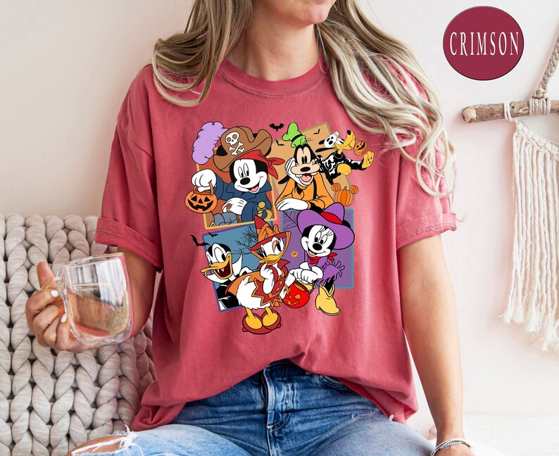 K&ouml;nnte beinhalten: Ein rotes T-Shirt mit einem Grafikdruck von Mickey Mouse, Minnie Mouse, Donald Duck, Goofy und Pluto in Halloween-Kost&uuml;men. Die Figuren halten S&uuml;&szlig;igkeiten und K&uuml;rbisse. Der Text "CRIMSON" ist in der oberen rechten Ecke des Bildes sichtbar.