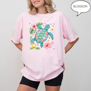 May include: A light pink t-shirt featuring a turquoise sea turtle design with floral accents. The turtle is surrounded by tropical flowers and leaves. The word "BLOSSOM" is in a speech bubble.