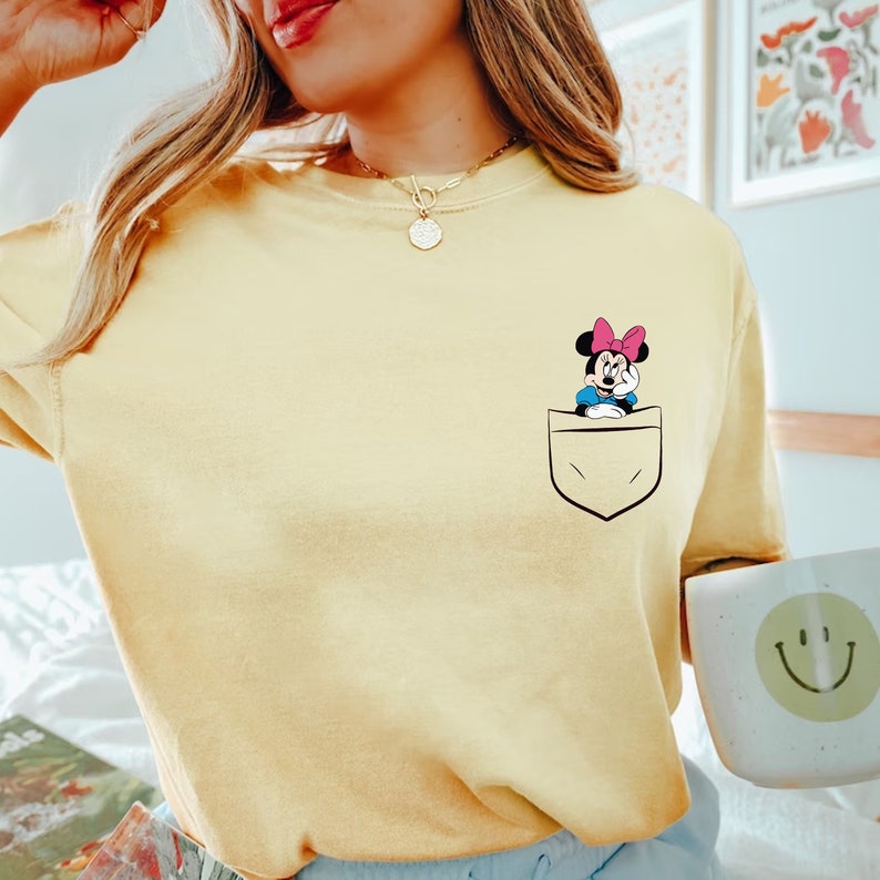May include: A yellow t-shirt with a pocket featuring a cartoon image of Minnie Mouse. Minnie Mouse is wearing a pink bow and is peeking out of the pocket.