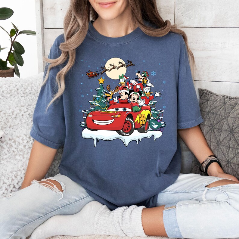 May include: A blue t-shirt with a Christmas-themed design featuring Mickey Mouse and friends riding in a red Lightning McQueen car. The design includes a Christmas tree, snowflakes, and a moon.