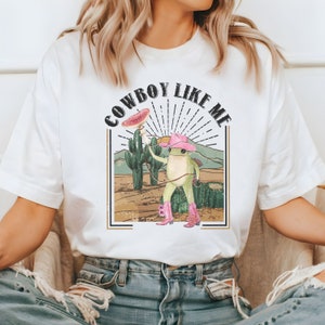 May include: White t-shirt with a graphic of a frog wearing a cowboy hat and boots, holding a slice of watermelon. The text "COWBOY LIKE ME" is printed above the frog.