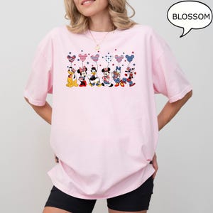 Puede incluir: Una camiseta rosa claro con un gr&aacute;fico de personajes de dibujos animados sosteniendo globos en forma de coraz&oacute;n con patrones rojos, blancos y azules. La palabra "BLOSSOM" est&aacute; en un bocadillo. La camiseta tiene mangas cortas.