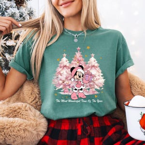 May include: A teal green t-shirt featuring a Christmas design. The graphic includes pink Christmas trees, stars, and a cartoon mouse holding a gift. The text reads "The Most Wonderful Time Of The Year."