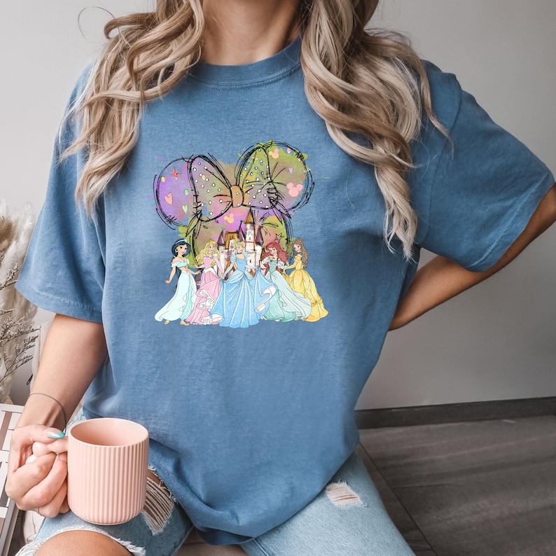 Puede incluir: Camiseta azul con un gr&aacute;fico colorido de princesas de Disney y un castillo. El dise&ntilde;o incluye un lazo y orejas de rat&oacute;n. Las princesas visten vestidos de varios colores, incluyendo azul, amarillo y rosa.