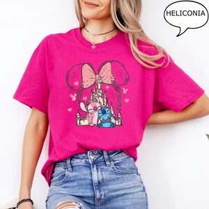 Puede incluir: Una camiseta magenta con un gr&aacute;fico de dos personajes de dibujos animados, uno rosa y otro azul, frente a un castillo. El dise&ntilde;o incluye un lazo rosa y orejas de rat&oacute;n. La palabra "HELICONIA" est&aacute; en un bocadillo.