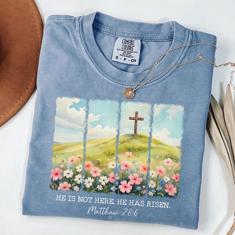 May include: A light blue t-shirt featuring a watercolour design of a cross on a hill with flowers. The text "HE IS NOT HERE. HE HAS RISEN. Matthew 28:6" is printed on the shirt.
