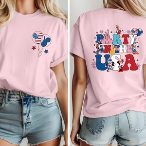 K&ouml;nnte beinhalten: Ein hellrosa T-Shirt mit einem patriotischen Design. Die Vorderseite zeigt Ballons mit amerikanischen Flaggenmustern und Sternen. Auf der R&uuml;ckseite des Shirts steht der Text "Party in the USA" mit Zeichentrickfiguren und einem Schloss.