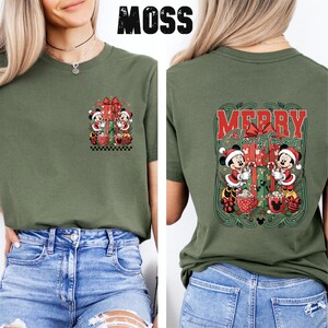 May include: Moss green t-shirt with a Christmas design featuring Mickey and Minnie Mouse. The front has a small graphic, and the back displays a larger "Merry Christmas" design with the characters. The word "MOSS" is printed at the top.