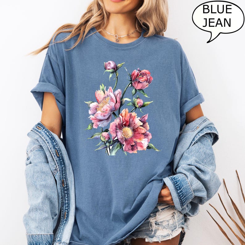 Pu&ograve; includere: T-shirt blu con un design floreale ad acquerello di peonie rosa e pesca e foglie verdi. Il testo "BLUE JEAN" &egrave; in un fumetto. La maglietta ha le maniche arrotolate.