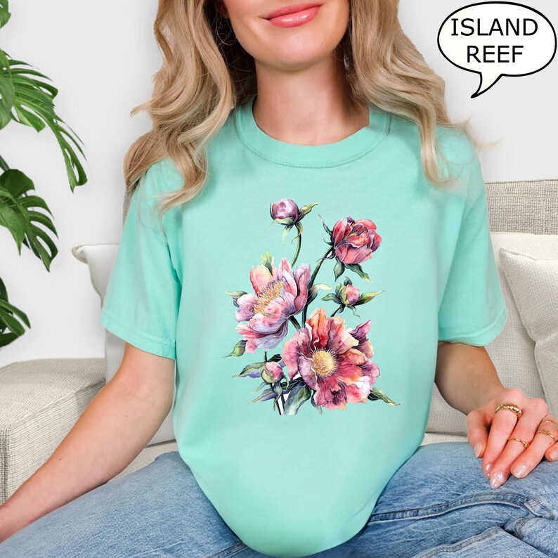 Pu&ograve; includere: Una maglietta turchese chiaro con un design floreale ad acquerello nei toni del rosa e del rosso. La grafica include peonie e boccioli. Il testo "ISLAND REEF" &egrave; in una nuvoletta.