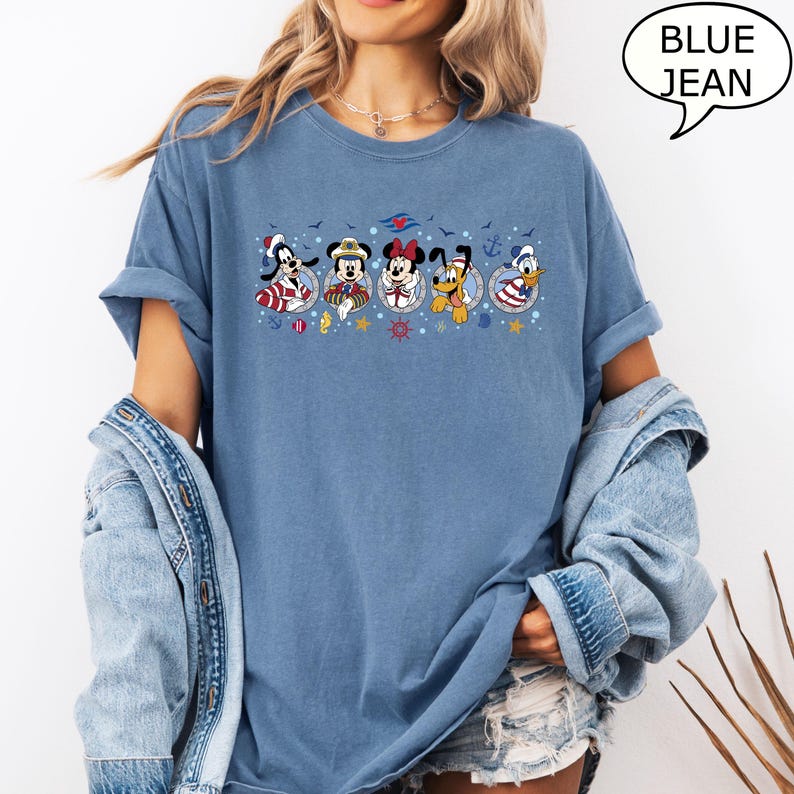 May include: Blue jean-colored t-shirt featuring a graphic of Disney characters in nautical attire. The characters are arranged in a row, each within a circular frame. The shirt has rolled-up sleeves and the text "BLUE JEAN" in a speech bubble.