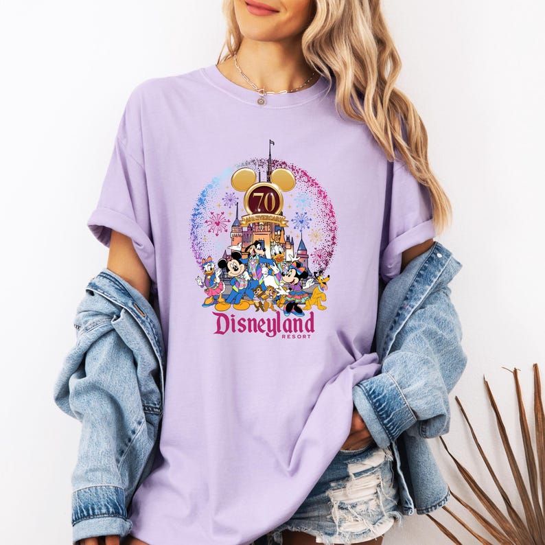 May include: A purple t-shirt with a graphic of Mickey Mouse and friends celebrating Disneyland's 70th anniversary. The graphic features the Disneyland castle, fireworks, and the text "Disneyland Resort".
