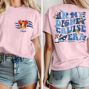 Op de afbeelding: Roze T-shirt met een cartoonhond met een kapiteinshoed op de voorkant en de tekst "In My Disney Cruise Era" met cartoonfiguren op de achterkant.