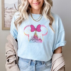 Puede incluir: Una camiseta azul claro con un lazo rosa y una ilustraci&oacute;n colorida de un castillo. El castillo est&aacute; rodeado por un contorno azul claro y amarillo.