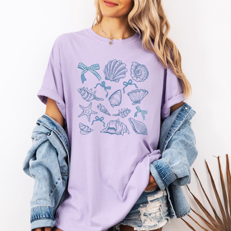 May include: A lavender t-shirt with a blue and white seashell pattern. The shirt has a relaxed fit and short sleeves.