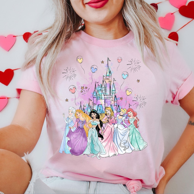 Puede incluir: Camiseta rosa con un gr&aacute;fico de princesas Disney de pie frente al castillo de Cenicienta con fuegos artificiales y globos.