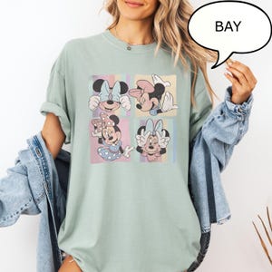 Puede incluir: Camiseta verde claro con un dise&ntilde;o de cuatro paneles de Minnie Mouse en varias poses. Cada panel tiene un fondo pastel con rayas verticales. La palabra "BAY" est&aacute; en una burbuja.