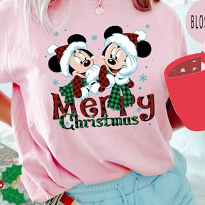 May include: Pink t-shirt with a Christmas design featuring Mickey and Minnie Mouse wearing Santa hats and scarves. The text "Merry Christmas" is written in red and black plaid.