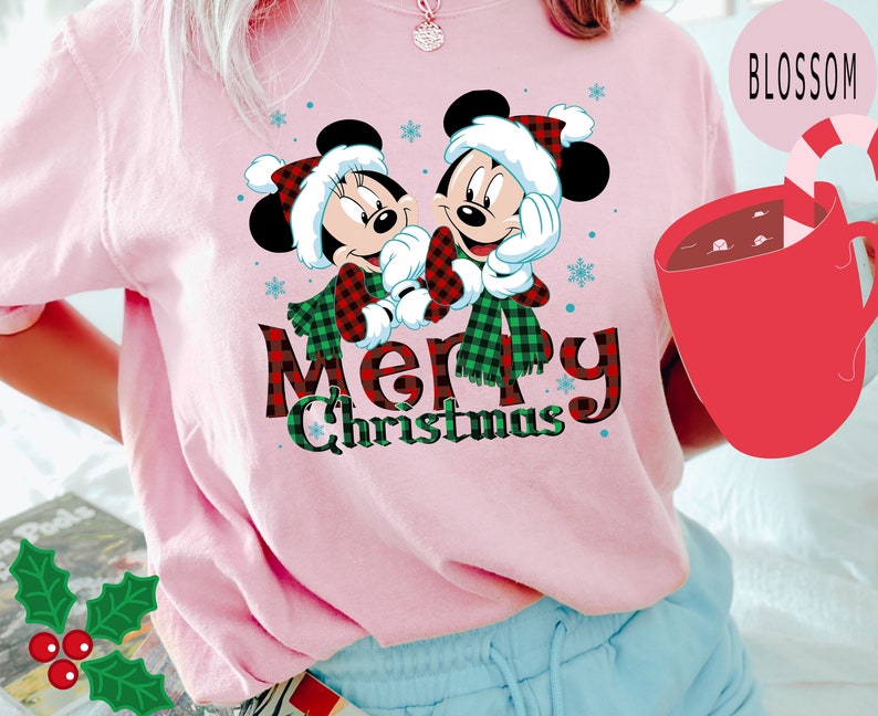 May include: Pink t-shirt with a Christmas design featuring Mickey and Minnie Mouse wearing Santa hats and scarves. The text "Merry Christmas" is written in red and black plaid.