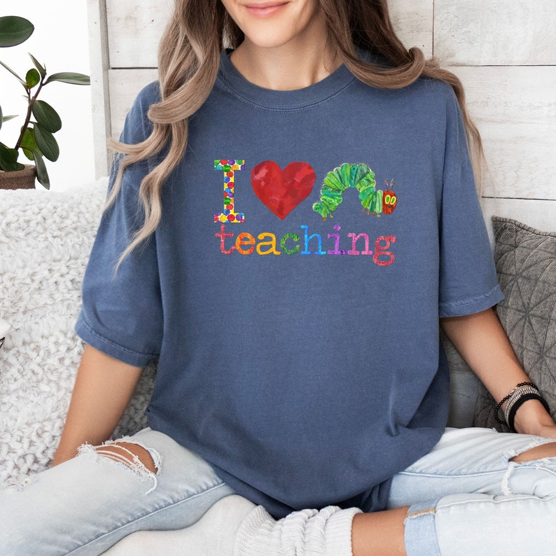 Op de afbeelding: Een blauw T-shirt met de tekst "Ik hou van lesgeven" in regenboogkleuren. Een rood hart en een groene rups bevinden zich in het midden van de tekst.