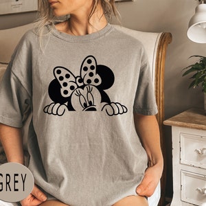 K&ouml;nnte beinhalten: Ein graues T-Shirt mit einem schwarz-wei&szlig;en Cartoon-Bild von Minnie Mouse, die &uuml;ber den oberen Rand schaut. Das Bild enth&auml;lt Minnies charakteristisches Band mit Punkten.